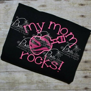 Pode incluir: Camiseta preta com bordado rosa que diz "My Mom Rocks!" com um violão rosa e branco com estampa de zebra em forma de coração.