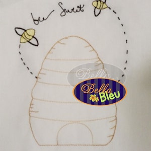 May include: Embroidered design of a beehive with the text "bee sweet" and three bees flying around it. The design is on a white fabric background.