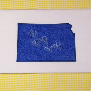 May include: A white t-shirt with a blue outline of the state of Kansas. The state is filled with a blue fabric with a white speckled pattern.