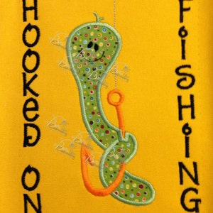 Könnte beinhalten: Ein grüner Wurm mit einem lächelnden Gesicht ist an einer Angelschnur befestigt. Der Text "HOOKED ON FISHING" ist auf gelbem Hintergrund gestickt.