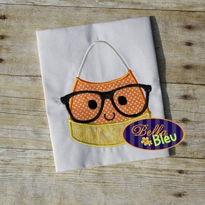 Może przedstawiać: Haftowany wzór przedstawiający kukurydzę cukrową w okularach na białym materiale. Kukurydza cukrowa jest pomarańczowa, żółta i biała z czarnymi okularami. Tekst "Bella Bleu" znajduje się w prawym dolnym rogu obrazu.