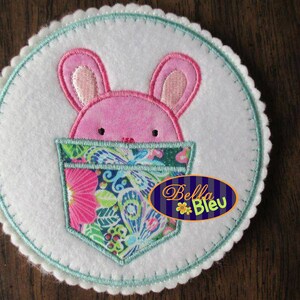 May include: A white felt applique with a pink bunny rabbit peeking out of a floral pocket. The text "Bella Bleu" is embroidered in blue on the felt.