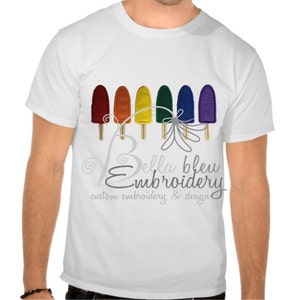 May include: White t-shirt featuring a rainbow popsicle design and the text "Bella Bleu Embroidery, custom embroidery & design".