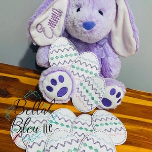 May include: Purple plush bunny with white ears and a white embroidered name "Emma" on one ear. The bunny is holding a set of 8 white felt Easter eggs with green, blue, and purple embroidered designs.