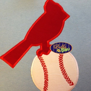 May include: A red cardinal bird applique perched on a baseball, with red stitching details. The design is set against a light blue background. The image includes a small oval tag with the text "Bella Bleu".