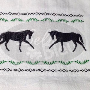 Puede incluir: Toalla de cocina blanca con caballos bordados en negro y enredaderas de hojas verdes. La toalla tiene un borde geométrico blanco y negro.