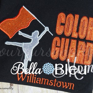 Puede incluir: Una camiseta negra con un diseño de purpurina naranja que dice "Color Guard" y una silueta plateada de purpurina de una bailarina de la guardia de color sosteniendo una bandera. El texto "Bella Bleum Williamstown" también está en la camiseta en purpurina plateada.