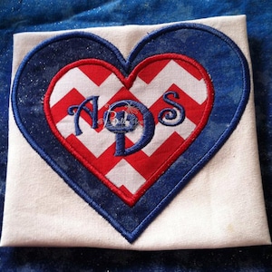 Op de afbeelding: Een witte stoffen hartvormige patch met een rood-wit chevron-patroon erin. Het chevron-patroon heeft een blauwe omtrek en de letters "ADS" zijn in blauw geborduurd op het chevron-patroon.