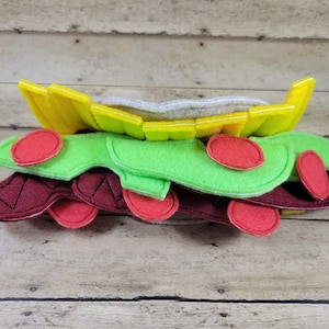 Puede incluir: Un juguete de fieltro en forma de sándwich con capas de lechuga, tomate y queso. La capa superior es un pan de fieltro amarillo con una parte superior de fieltro blanco.