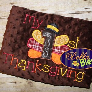 以下が含まれることがあります： オレンジ、赤、黄色の糸で「my 1st Thanksgiving」と刺繍された茶色のぬいぐるみ毛布。茶色のフェルトの数字1に黄色の帽子がついたものが毛布の中央にあります。「Bella Bleu」という文字が、黄色の花がついた青い楕円形に刺繍されています。