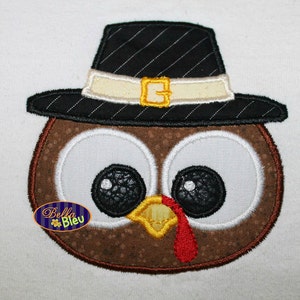 以下が含まれることがあります： 茶色の七面鳥の刺繍アップリケ。大きな白い目、黄色いクチバシ、赤い肉垂れがあります。七面鳥は、白いバンドと金色のバックルが付いた黒い巡礼者の帽子をかぶっています。