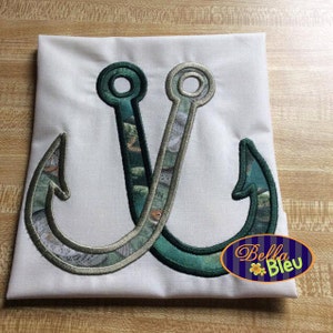 以下が含まれることがあります： 魚柄の緑と白の刺繍が施された釣り針が2つ。針は白い布地に縫い付けられています。