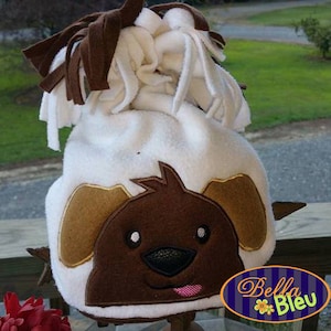 Può includere: Cappello in pile bianco con un'applicazione in feltro marrone e beige del viso di un cane. Il cappello ha lacci in pile marrone sulla parte superiore. Il cappello è appoggiato su un tavolo con un fiore rosso in primo piano. Il testo "Bella Bleu" è visibile sullo sfondo.