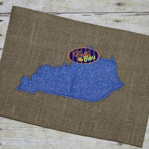 Könnte beinhalten: Eine blaue gestickte Umrandung des Bundesstaates Kentucky auf einem braunen Jute-Hintergrund. Der Text "Bella Bleu" ist in einem weißen Oval mit gelber Umrandung gestickt.