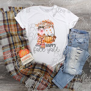 May include: White t-shirt with a graphic of a pig wearing a scarf and a hat, sitting under a tree with fall leaves. The text "HAPPY fall y'all" is below the pig.