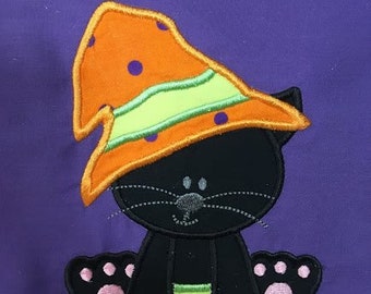 Puede incluir: Un gato negro con un sombrero de bruja naranja y verde con lunares negros. El gato está sentado con las patas extendidas hacia adelante.