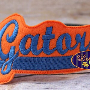 May include: Orange felt headband with blue embroidered text "Gators" and a blue stripe. The headband is decorated with a small blue and white flower logo that says "Bella Bleu".