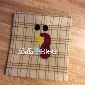 Può includere: Un tessuto a quadri beige e marrone con un disegno di tacchino ricamato. Il tacchino ha la testa rossa, il becco giallo e gli occhi neri. Il testo "Bella Bleu" è ricamato sotto il tacchino.