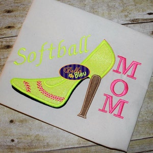 May include: A white fabric with the words "Softball Mom" embroidered in pink and yellow. The word "Mom" is in large letters and the word "Softball" is above the word "Mom". A yellow high-heeled shoe with a pink baseball stitch design is embroidered to the left of the words.