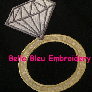 Puede incluir: Un anillo de oro con un gran diamante en la parte superior. El anillo está decorado con un patrón floral blanco. El texto "Bella Bleu Embroidery" es visible debajo del anillo.