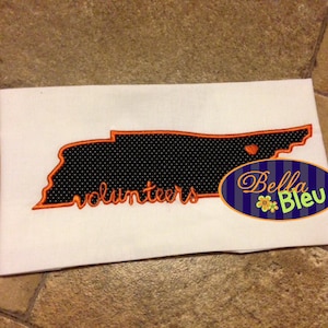 Könnte beinhalten: Ein weißes T-Shirt mit einer orangefarbenen Stickerei des Bundesstaates Tennessee. Die Umrandung ist mit schwarzen Punkten gefüllt und das Wort "Volunteers" ist in Orange unterhalb der Umrandung des Bundesstaates gestickt. Das T-Shirt ist eine leere Leinwand für ein individuelles Design.