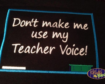May include: A black chalkboard with a blue frame and white text that reads "Don't make me use my Teacher Voice!" There is a white chalk line and a green ledge at the bottom of the chalkboard.
