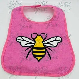 Peut inclure: Bavoir rose pour bébé avec un motif d'abeille. Le bavoir a une forme arrondie, une bordure rose et une fermeture à bouton-pression. L'abeille est jaune et noire avec des ailes blanches.