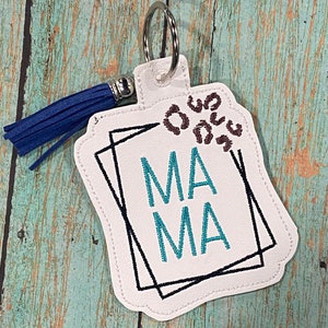 May include: White fabric keychain with a blue tassel. The keychain has a black and white geometric design with the word "MAMA" in teal lettering. The keychain has a brown leopard print design in the top corner.