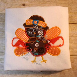 以下が含まれることがあります： 茶色とオレンジ色の刺繍された七面鳥で、茶色の帽子と青いリボンが付いています。七面鳥は模様のある体と黄色いくちばしをしています。