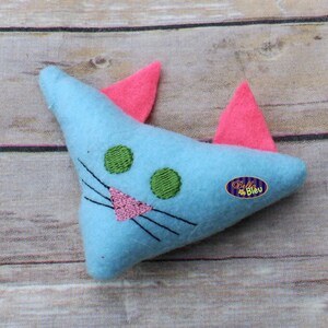 Puede incluir: Un juguete de gato de fieltro azul claro con orejas rosas y ojos verdes. El juguete tiene una nariz rosa y bigotes negros.