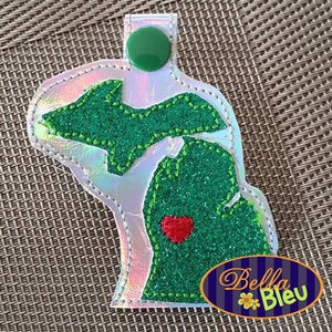 Puede incluir: Un llavero blanco iridiscente con un contorno de fieltro verde del estado de Michigan. El estado tiene un corazón rojo en el centro y el texto "Bella Bleu" está impreso en la parte inferior.