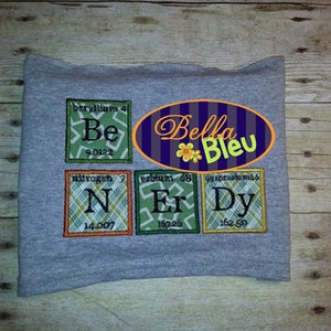 May include: A gray sweatshirt with a green, orange, and white patterned design featuring the chemical symbols for Beryllium, Nitrogen, Erbium, and Dysprosium. The text "Bella Bleu" is in a purple oval with a white flower.