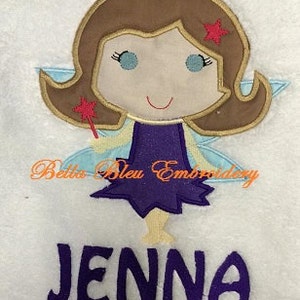 Peut inclure: Une broderie représentant une fée aux cheveux bruns, vêtue d'une robe violette et d'ailes bleues. La fée tient une baguette rose avec une étoile. Le nom "Jenna" est brodé en dessous. Le texte "Betta Bleu Embroidery" est également visible.