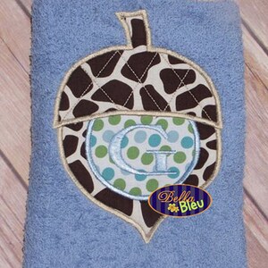 以下が含まれることがあります： 茶色と白の刺繍が施された青いタオル。どんぐりには緑と青のドット模様の中心に白いGの文字が刺繍されています。