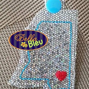 May include: A silver glitter key chain with a blue circle and a red heart. The key chain is shaped like the state of Alabama and has the text "Bella Bleu" on it.
