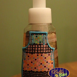 May include: A clear plastic bottle with a white pump dispenser and a colourful fabric apron design. The bottle is labelled "8.75 fl oz / 259 ml" and has a blue and white label that says "Bella Bleu".