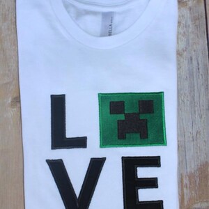 May include: White t-shirt with the word "LOVE" in black outlined letters. The "O" is replaced with a green square containing a black Minecraft Creeper face.