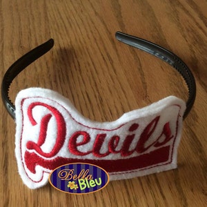 Puede incluir: Una diadema blanca con un parche de fieltro rojo que dice "Devils" en letra cursiva. La diadema es negra y tiene un diseño simple.