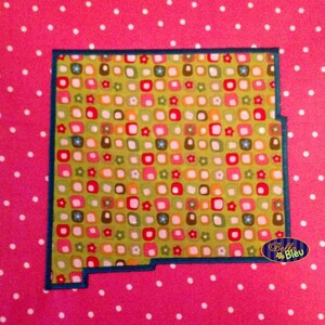 May include: A green fabric with a pattern of colorful squares and flowers, outlined in dark blue, in the shape of the state of New Mexico. The fabric is on a pink background with white polka dots. The text "Bella Bleu" is in the lower right corner of the image.