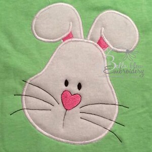 May include: White bunny rabbit face with pink nose and black eyes on a green background.