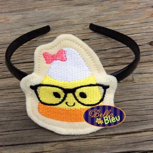 May include: A yellow and orange candy corn with a white top and a pink bow, wearing black glasses and a happy face, is attached to a black headband. The headband is on a wooden surface.