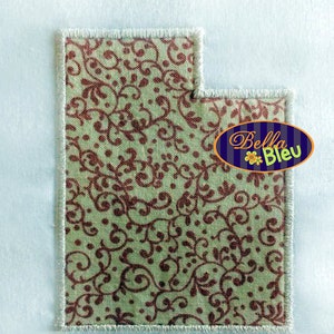 May include: A green fabric with a brown floral pattern, with a white stitched border. The fabric is shaped like the state of Utah. The text "Bella Bleu" is in a blue oval with a yellow flower.