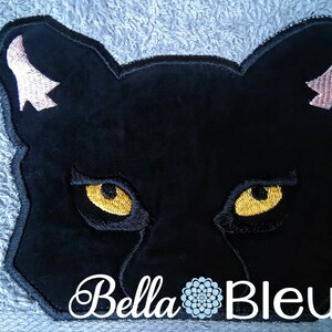 Peut inclure: Visage de panthère noire brodé avec des oreilles roses et des yeux jaunes sur un fond bleu. Le texte "Bella Bleu" est sous la panthère.