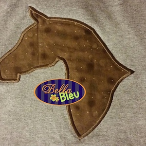 May include: A brown horse head silhouette embroidered on a gray sweatshirt. The silhouette is outlined in brown thread and has a speckled brown fabric inside. The text "Bella Bleu" is visible in a blue oval with a white flower.