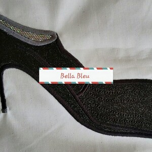 May include: Black sparkly fabric high heel shoe applique with a gray trim and a silver sequin detail. The applique is on a white fabric background. The text "Bella Bleu" is on a red and blue striped banner.