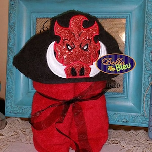 May include: A red hooded towel with a black embroidered boar head with white tusks. The towel is tied with a black ribbon and is displayed in a blue frame. The text "Bella Bleu" is visible in the image.