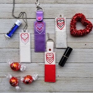 May include: Five different lip balm holders featuring a heart design. The holders are made from various materials, including leather, fabric, and plastic. The holders come in a range of colours, such as purple, white, red, and black. Each holder is adorned with a heart design.