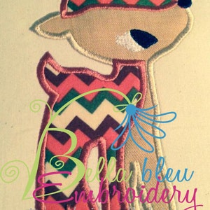 May include: Embroidered applique of a deer with a chevron patterned hat and body. The deer is pink, green, and white. The text "Bella bleu embroidery" is visible in the bottom right corner.