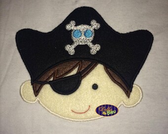 May include: Embroidered applique of a cartoon pirate with a black eye patch, brown hair, and a black pirate hat with a white skull and crossbones design. The pirate is smiling.