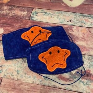 May include: Two blue fabric face masks with orange star-shaped embroidery. The stars have a smiley face with a frown.